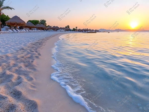 A serene sandy beach at sunset, the image shows calm blue sea water gently lapping against the shore. On the beach, there are straw parasols and palm trees lining the coastline. The sun rises on the horizon, creating a warm golden glow in the sky and on the water. The scene appears picturesque and tranquil.