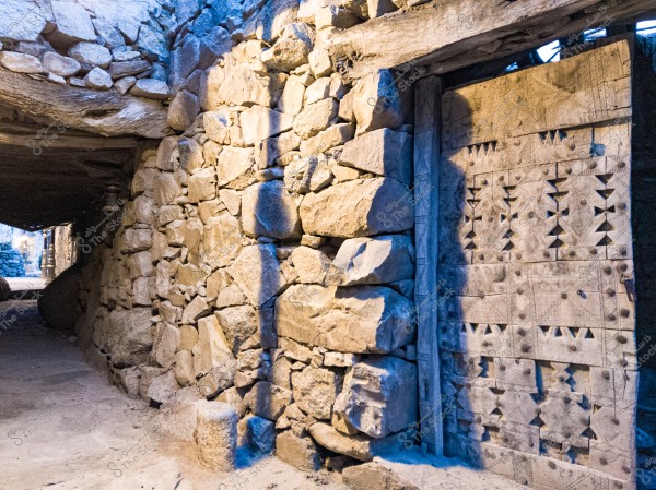 A scene of an old stone wall made from irregular stones, connected to a wooden door decorated with geometric patterns. Light shines on the wall, highlighting its details. In the background, a dark passageway creates a contrast with the light illuminating the stone surface and door.