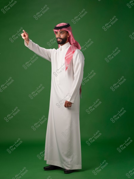 The image shows a man standing against a green background, wearing a white thobe and a traditional Saudi headscarf. He is pointing forward with his hand and appears to be smiling. The scene conveys confidence and a positive expression.
