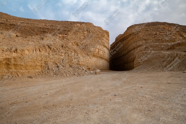 An image of a split rock cliff with a narrow passage between the two halves. The cliff is composed of layers of brown sandstone, and the dirt road in front appears empty of any vegetation or people. The sky in the background is cloudy with light clouds.
