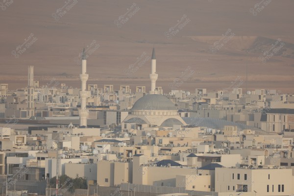 A view of a modern city with numerous clustered buildings, featuring a mosque in the center with a large dome and two tall minarets. The area surrounding the buildings is desert-like, barren, and appears distant from the city.