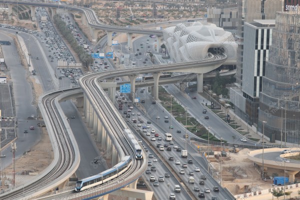 An image of a city depicting a modern urban scene in the Middle East, featuring an elevated metro line running above highways. The picture shows numerous cars driving along the congested road with multi-level roadways. In the background, a distinctive architectural building is visible with a modern and contemporary design.