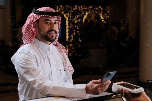 شاب سعودي في استقبال الفندق ، ويقوم بتعابير مختلفة باستخدام الجوال  ، ويقوم بدفع عبر جهاز الدفع الالكتروني