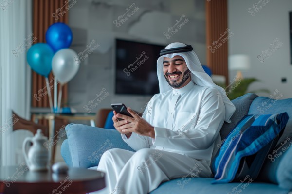 A portrait of a man sitting on a blue sofa in a living room, wearing a traditional white Arabic thobe with a headscarf (keffiyeh and agal). He is smiling and looking at his mobile phone. In the background, there are blue and white balloons, a table with a traditional coffee pot, and a blurred wall with a TV.