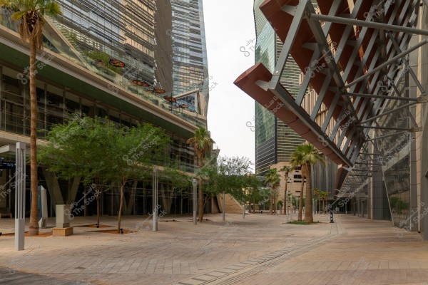 تظهر الصورة مشهدًا حضريًا حديثًا مع مباني زجاجية مرتفعة ومصممة بتصميمات معمارية مبتكرة. تحيط المباني بمجال مفتوح مزين بالنخيل وأشجار صغيرة. الأرضية مبلطة بألواح حجرية, وتوجد رفاصات معدنية كبيرة على مواجهة الجدران.