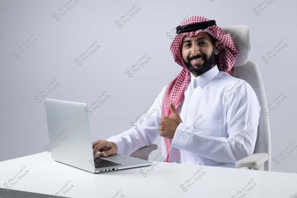 An image of a man sitting in front of a laptop on a white desk. He is wearing a white thobe and a red and white checkered ghutra with a black agal, giving a thumbs-up with a smile. The background is light gray.