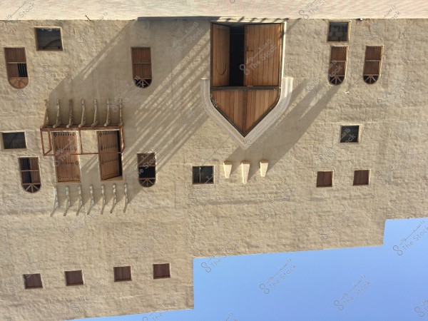Image of a facade of a traditional building with ancient Arabic architectural style. The facade is made of clay or stone, featuring rectangular wooden windows and some arched ones. There is an ornate wooden projecting balcony, with shadows casting distinct shapes on the wall. The sky is clear and blue.