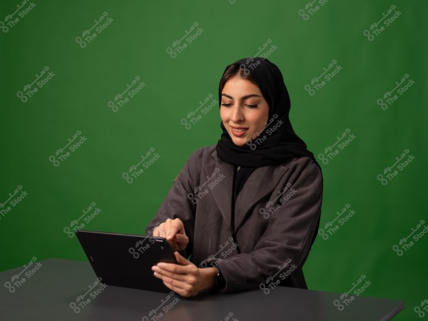 امرأة ترتدي حجابًا تجلس على مكتب وتستخدم جهازًا لوحيًا بخلفية خضراء اللون.