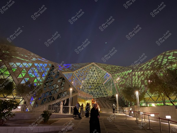 A photo of a modern architectural building featuring geometric structures made up of triangles illuminated with multiple colors, including green and blue. The photo is taken at night with a clear night sky. In the foreground, several people are walking or sitting near the building, surrounded by green plants and lit lamps.