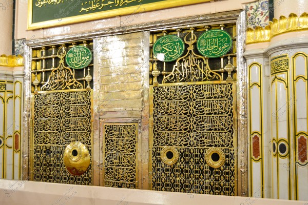 The image shows the golden-plated metal grille covering the Rawdah Ash-Sharifah in Al-Masjid an-Nabawi in Medina. The architectural design features prominent brass details and Islamic patterns, with beautiful Arabic script inscriptions displayed on the surface. Circular plaques above the grille in green display the names of the companions.