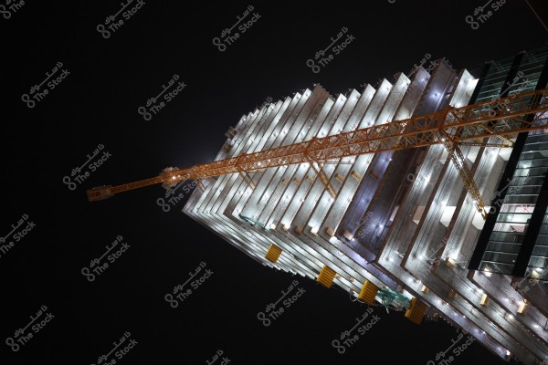 Image of a building under construction at night, illuminated with white lights. An orange crane extends across the multi-story structure. The sky in the background is dark.