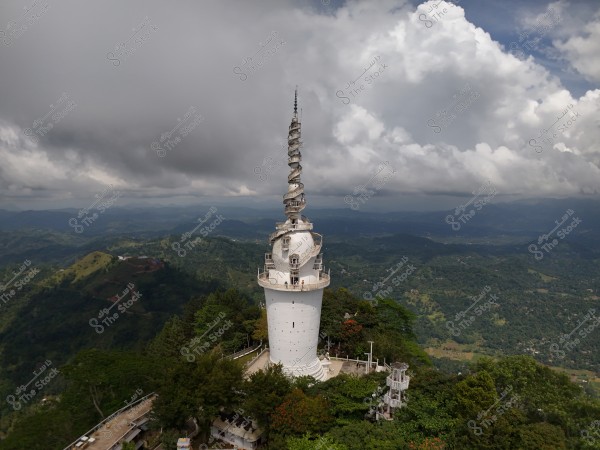 Image of a tall white tower situated amidst dense green forests on a mountaintop. The tower features a unique architectural design with a metal spiral at the top. Dark clouds cover the sky, adding a dramatic feel to the scene. Hills extend into the distance towards the horizon.
