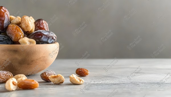 An image of a wooden bowl filled with dates and nuts, placed on a smooth gray surface. The background features a blurred gray wall. Some dates and nuts are scattered beside the bowl.