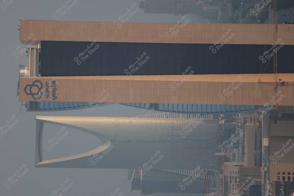 The image shows a large skyscraper belonging to Al Rajhi Bank displayed horizontally, with the Kingdom Tower in Riyadh, Saudi Arabia, in the background. The top floor features a dark glass facade with a beige external structure. The horizon displays a view of the city with other buildings and residential complexes, and an orange crane truck on the right.