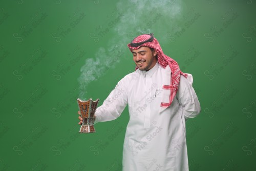 رجل سعودي يرتدي الزي التقليدي على خلفية خضراء ، التطيب بالبخور والعود ، استخدام خشب العود في المناسبات والأعياد.