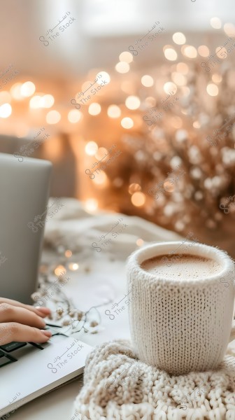 صورة لكوب مشروب ساخن مغطى بقماش محبوك بلون كريمي وبجانبه يد على لوحة مفاتيح جهاز كمبيوتر محمول. تظهر الخلفية بها أضواء خافتة صغيرة مما يضفي جواً دافئاً ومريحاً.