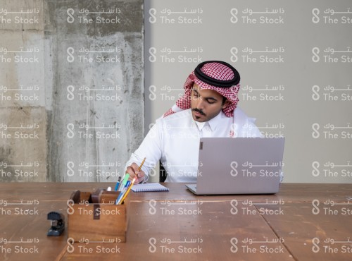 رجل سعودي يرتدي الزي السعودي التقليدي يجلس على طاولة الاجتماعات ويظهر أمامه الابتوب ومستلزمات مكتبية بينما يدون الملاحظات في الدفتر