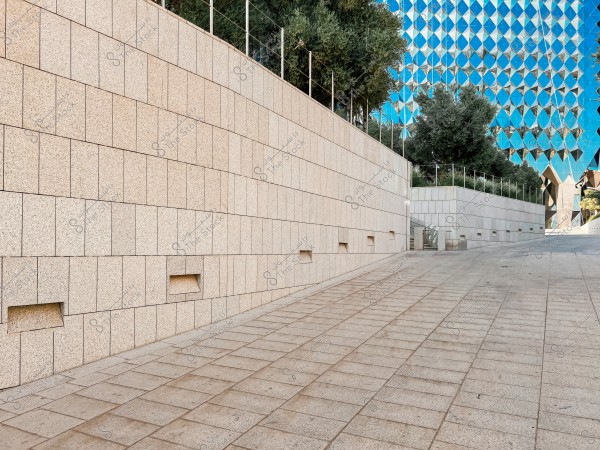 The image shows a long stone wall extending along a tiled pathway leading to a building with blue glass and a distinctive geometric pattern. Green trees overlook the wall, and the area appears calm and empty.