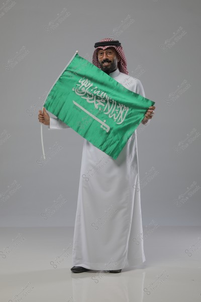 Image of a person wearing traditional Saudi attire, including a red and white shemagh on their head. The person is holding the green Saudi flag, which features the shahada and a sword. The person is smiling, with a light gray background.