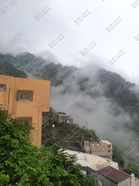 Image of a scenic view featuring a mountainous area covered with mist, creating a mysterious atmosphere. In the foreground, there is a building with a yellow facade and mesh windows, surrounded by lush green foliage. A cluster of other houses can be seen built on terraced hills in the distance.