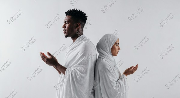 An image of a man and a woman wearing white Ihram clothing standing back-to-back. The man is on the left and the woman is on the right, both raising their hands in prayer. The scene is minimalistic with a neutral gray background.