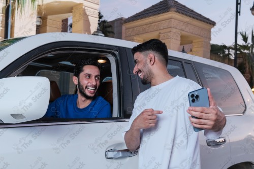 A snapshot of two young Saudis chatting and having fun on the road, their features showing joy and happiness, taking a selfie with a mobile phone