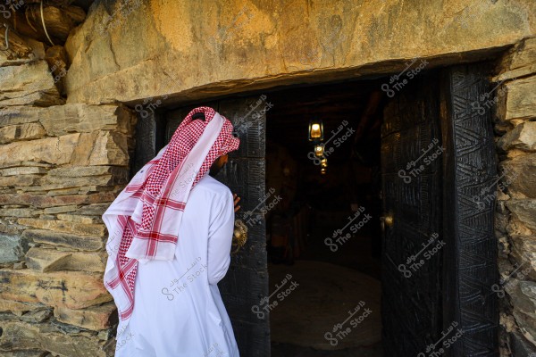 The image shows a person wearing traditional Arabic attire, including a white thobe and a red checkered headscarf, standing in front of an ornate wooden door. The person is opening the door and entering a space illuminated by traditional lanterns. The surrounding wall is made of natural stones.