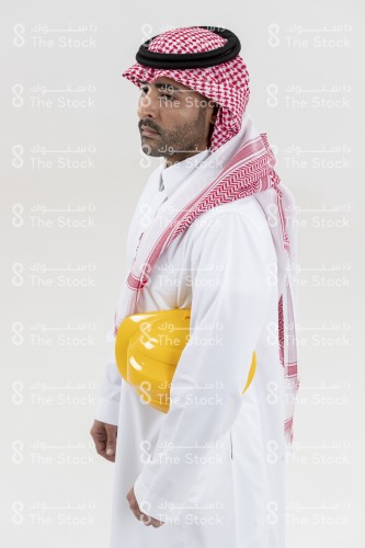 بورتريه صورة مقربة من الجانب لمهندس سعودي يرتدي الزي السعودي ويحمل تحت ذراعه خوذة المهندسين على خلفية بيضاء.