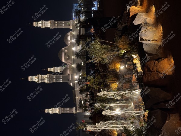 صورة رائعة لمسجد مضاء في الليل, مع مآذن مرتفعة وواجهة مضاءة بشكل جميل. في المقدمة, تظهر حديقة صغيرة بها نباتات وصخور مضاءة بأضواء صغيرة, تضيف لمسة جمالية للمشهد الليلي الهادئ.