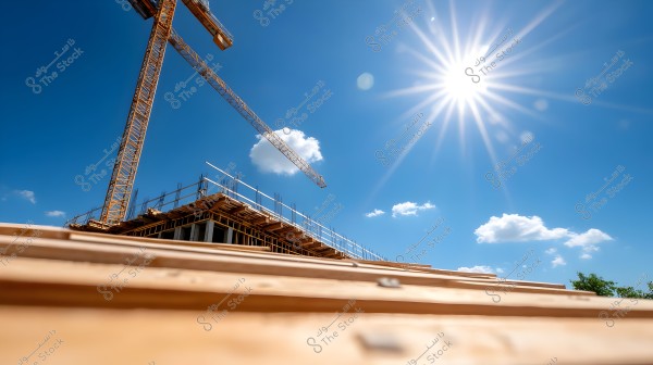 Image of a construction site featuring a large crane next to a building structure under construction, with a bright blue sky and some small clouds. The sun shines brightly in the upper right corner, with rays extending in all directions, and some trees visible in the bottom right of the image.