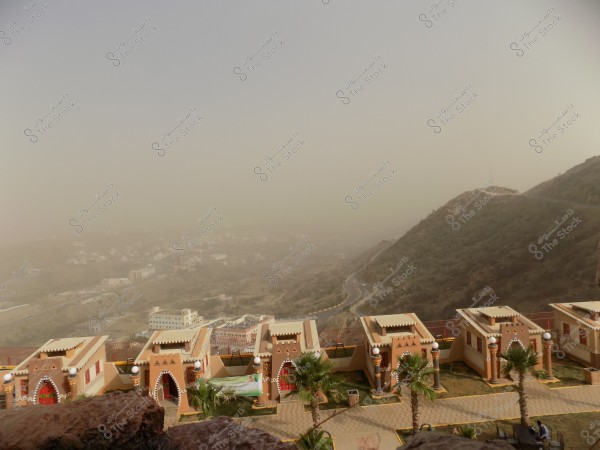 A panoramic view showing hills covered with vegetation, featuring traditional houses with light colors and decorative designs. Small palm trees are in front of the houses in the foreground, while a winding road runs through the hills. The atmosphere is cloudy with a hazy sky in the background.