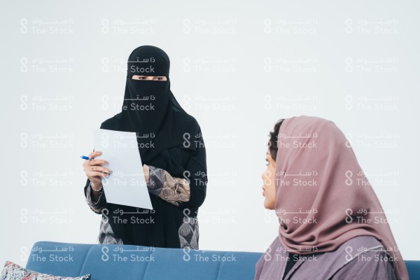 امرأة سعودية محجبة جالسة تلتفت لزميلتها التي تقف خلف الأريكة ترتدي العباءة والنقاب وتحمل بيديها الورقة والقلم أثناء نقاشهما