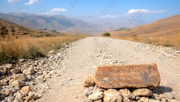 A dirt road extends through a dry mountainous landscape. Mountains surround the road in the distance, with some light white clouds in the clear blue sky. A stone marker on the side of the road features some inscriptions, and gravel is scattered on both sides of the road.