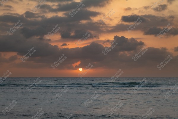 Sunset over a calm sea, with the orange sun touching the horizon line. Dark clouds obscure parts of the sky, with colors grading from orange to gray.