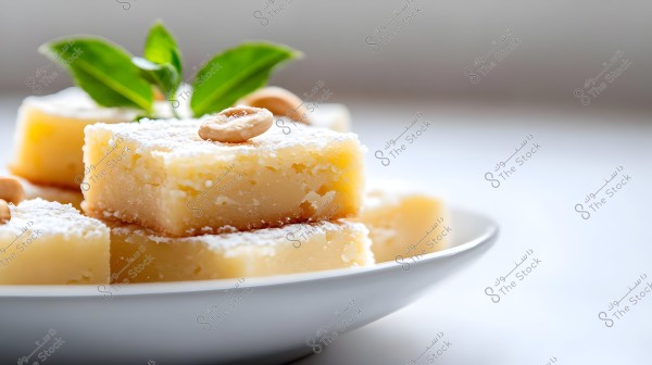 An image showing a collection of square-shaped sweets on a white plate. The sweets are dusted with powdered sugar and garnished with cashews and vibrant green leaves.