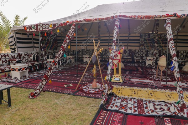 An image of a traditional tent adorned with intricate Arabic patterns in a desert setting. The tent features red carpets and furniture decorated in multiple colors, like black, white, and red. Inside the tent are seating areas decorated with geometric patterns, along with side tables and decorative vases. In the foreground, there is a wooden stand for a lance and traditional pottery.