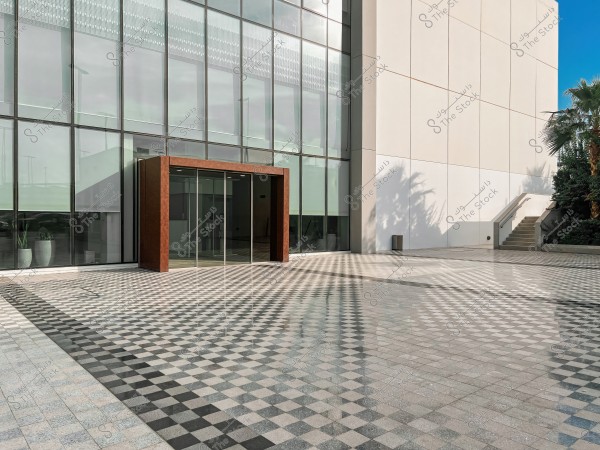 Entrance of a modern building with a large glass facade and a brown frame around the sliding glass doors. The floor is covered with square tiles in an interlocking pattern with white, gray, and black colors. There are some potted plants on the left, stairs on the right leading upward, and shadows of palm trees on the walls.