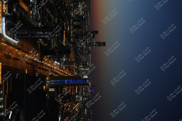 Night view of a large city featuring several skyscrapers and streetlights illuminating the roads. The sky in the background transitions from dark blue to red near the horizon. A tall tower adorned with blue lights is prominently featured in the center, acting as a focal point of the image.