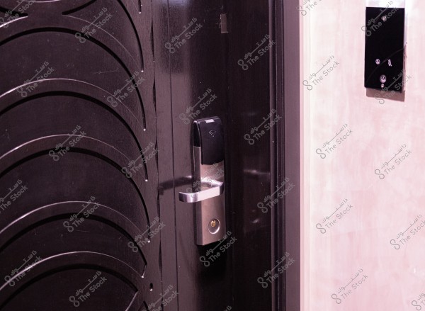 يظهر في الصورة باب أملس بلون داكن مزود بقفل إلكتروني حديث. يوجد على القفل قارئ بطاقة مغناطيسي ومقبض معدني بلون فضي. الجدار المجاور لباب يبدو بلون فاتح وله ملمس ناعم.