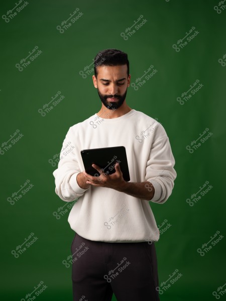 A portrait of a man standing against a green background, wearing a white sweater and dark pants, holding a tablet in his hands. He appears focused, looking down at the tablet.
