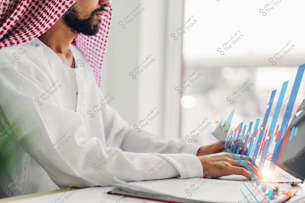 Image of a man wearing a white thobe and a red and white checkered headscarf, sitting in front of a laptop. Transparent graphs are overlaid on the image, suggesting data analysis or financial activities. The background is blurred.