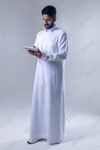 صورة لرجل يقف ويرتدي ثوبًا أبيض اللون, يحمل جهازًا لوحيًا بيديه. يبدو أنه من دول الخليج حيث يرتدي ثوبًا تقليديًا. الخلفية بيضاء وخالية من التفاصيل.