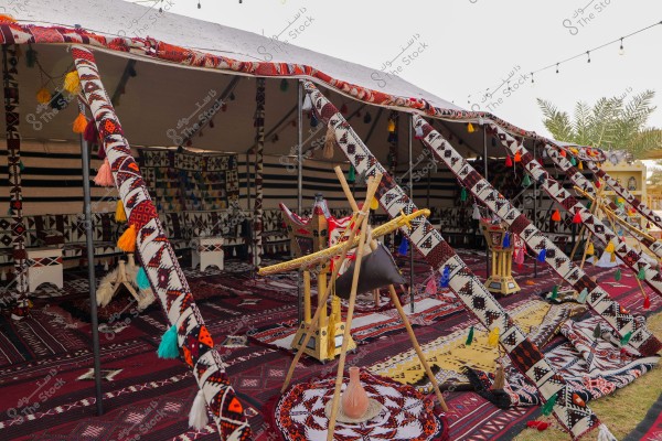 The image shows a traditional Bedouin tent adorned with colorful and varied decorations. Inside the tent, pillows and local-style furniture are visible, with distinct carpets covering the floor. The tent is decorated with colorful tassels and overhead hanging lights. The surroundings include palm trees, suggesting an eastern setting.