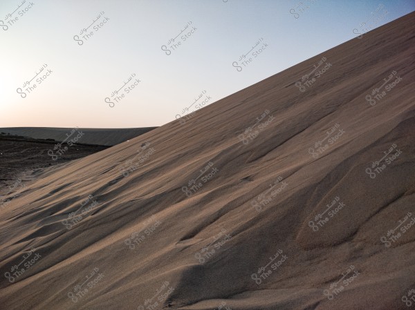 An image of a natural landscape featuring large, sloping sand dunes in the desert. The light reflects off the sand, highlighting the fine lines and details of the dunes. In the background, the horizon is clear, with the soft gradients of sunset or sunrise illuminating the sky.