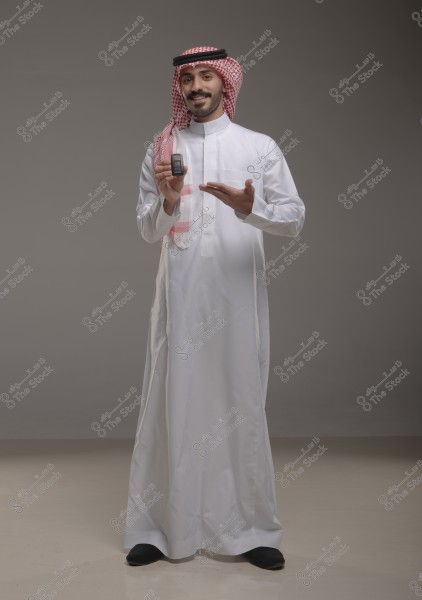 An image of a man wearing a white thobe standing against a neutral background. The man is holding a small device in one hand and pointing to it with the other. He is wearing a red and white ghutra and a black agal.