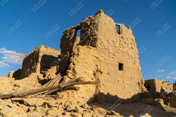 صورة تظهر أنقاض مبنى قديم مصنوع من الطين والطوب في منطقة صحراوية. يظهر المبنى جزئيًا منهدمًا وتحيط به قطع من الخشب المتساقطة. السماء زرقاء وصافية, مما يوحي بأجواء مشمسة.