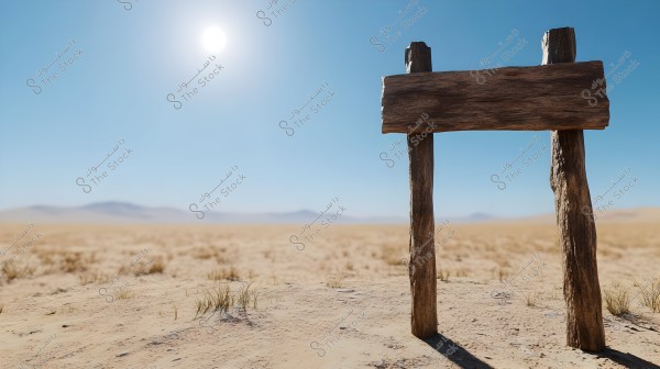 A vast desert under a clear blue sky with a bright sun. In the foreground, there is a simple wooden sign made of a horizontal plank supported by a frame of two vertical wooden posts, with no text on it. The landscape is filled with sand, with some dry vegetation scattered around, and distant hills visible on the horizon.