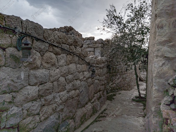 A narrow passageway made of old carved stones is surrounded by high stone walls with small metal lanterns connected by wires. A tree grows between the walls, and the sky is cloudy in the background.