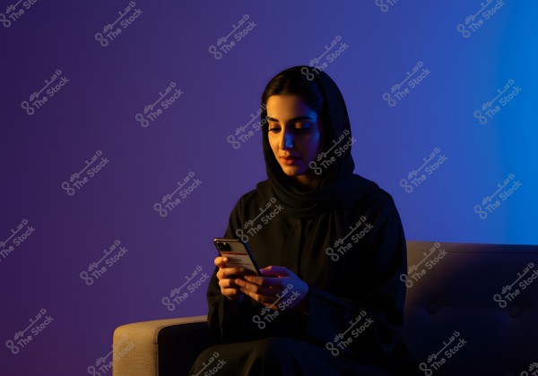A portrait of a woman wearing a black abaya and hijab, sitting on a sofa and using a smartphone. The background lighting in purple and blue gives the image a modern and stylish atmosphere. The clothing suggests she may be from a Gulf country like Saudi Arabia.