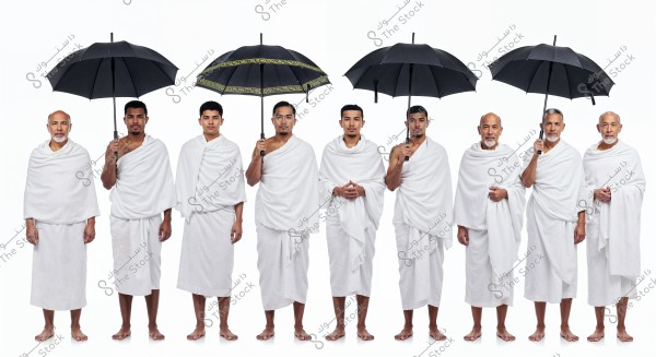 The image shows eight men standing in a straight line, wearing white Ihram garments, which are specific attire for performing Hajj or Umrah rituals. Some of them are holding black umbrellas. The men stand side by side in a single row, barefoot, indicating their readiness for religious practices in Saudi Arabia.